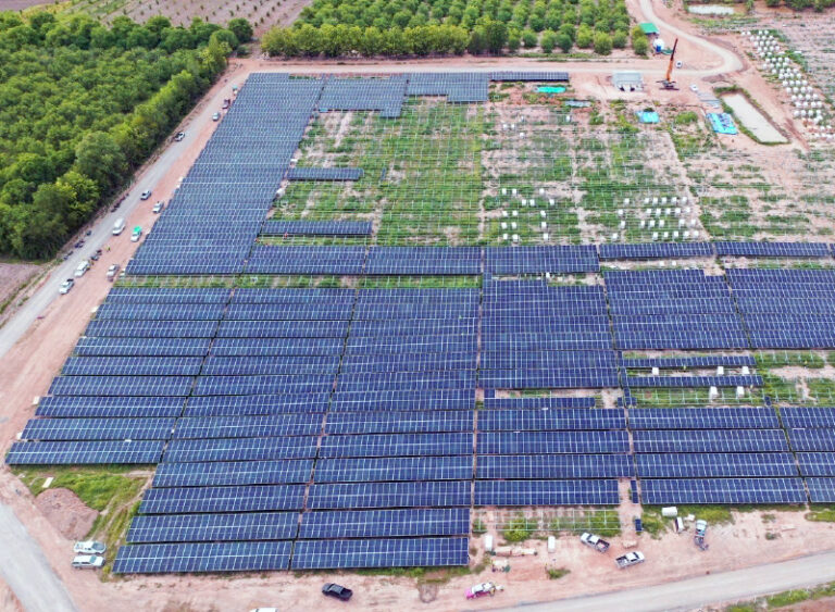 Aerial view of a large solar farm with rows of solar panels, surrounded by greenery and infrastructure.