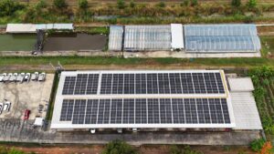 Aerial view of a solar panel installation on a large building, with greenhouses and ponds nearby.